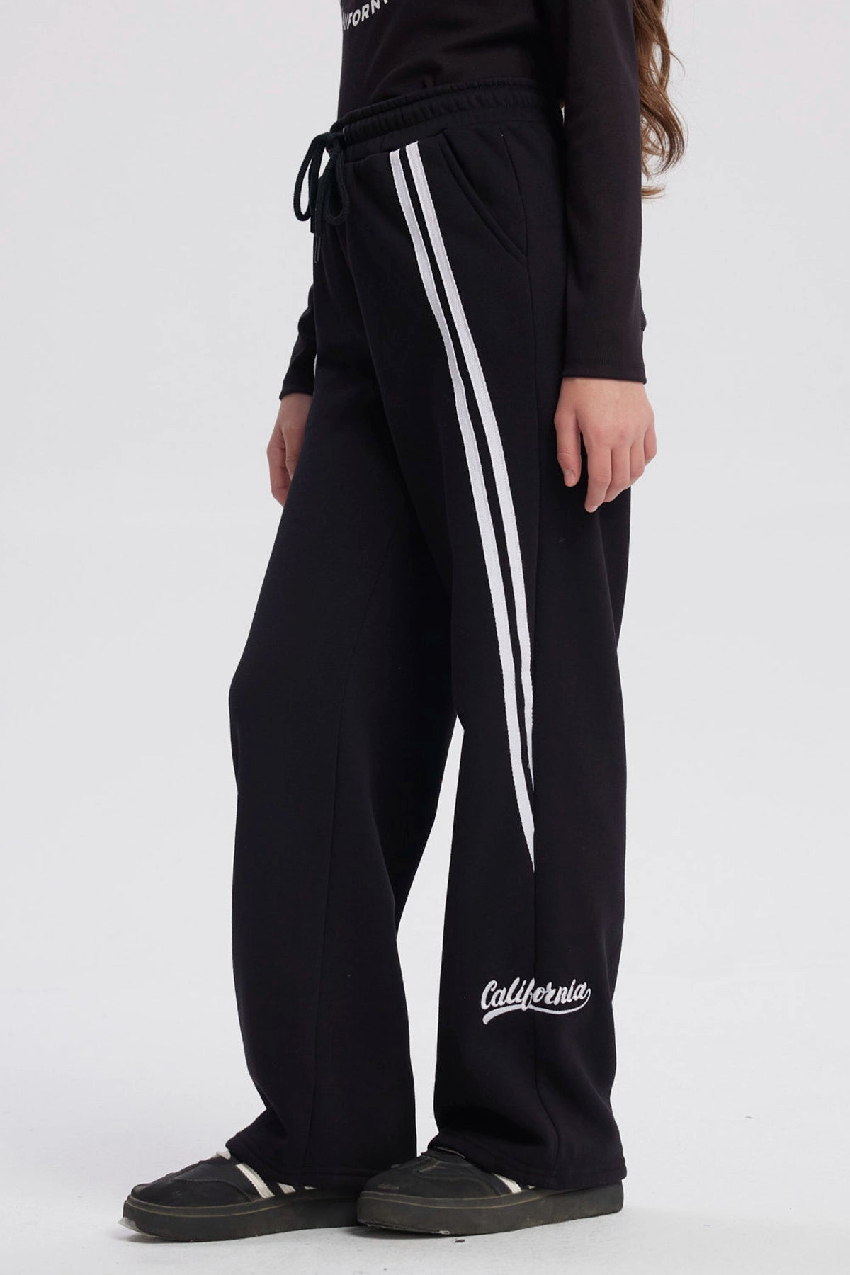 Pantalón buzo niña negro con corte wide leg líneas blancas verticales a los lados diseño deportivo y texto "active" en el tobillo