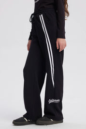 Pantalón buzo niña negro con corte wide leg líneas blancas verticales a los lados diseño deportivo y texto "active" en el tobillo