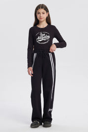 Pantalón buzo niña negro con corte wide leg líneas blancas verticales a los lados diseño deportivo y texto "active" en el tobillo
