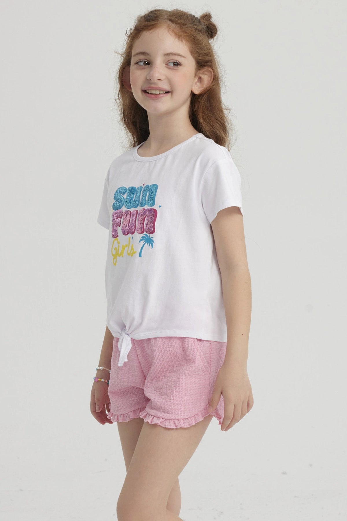 Polera blanca de niña con nudo frontal y estampado colorido que dice Sun Fun Girls con palmera pequeña al lado derecho
