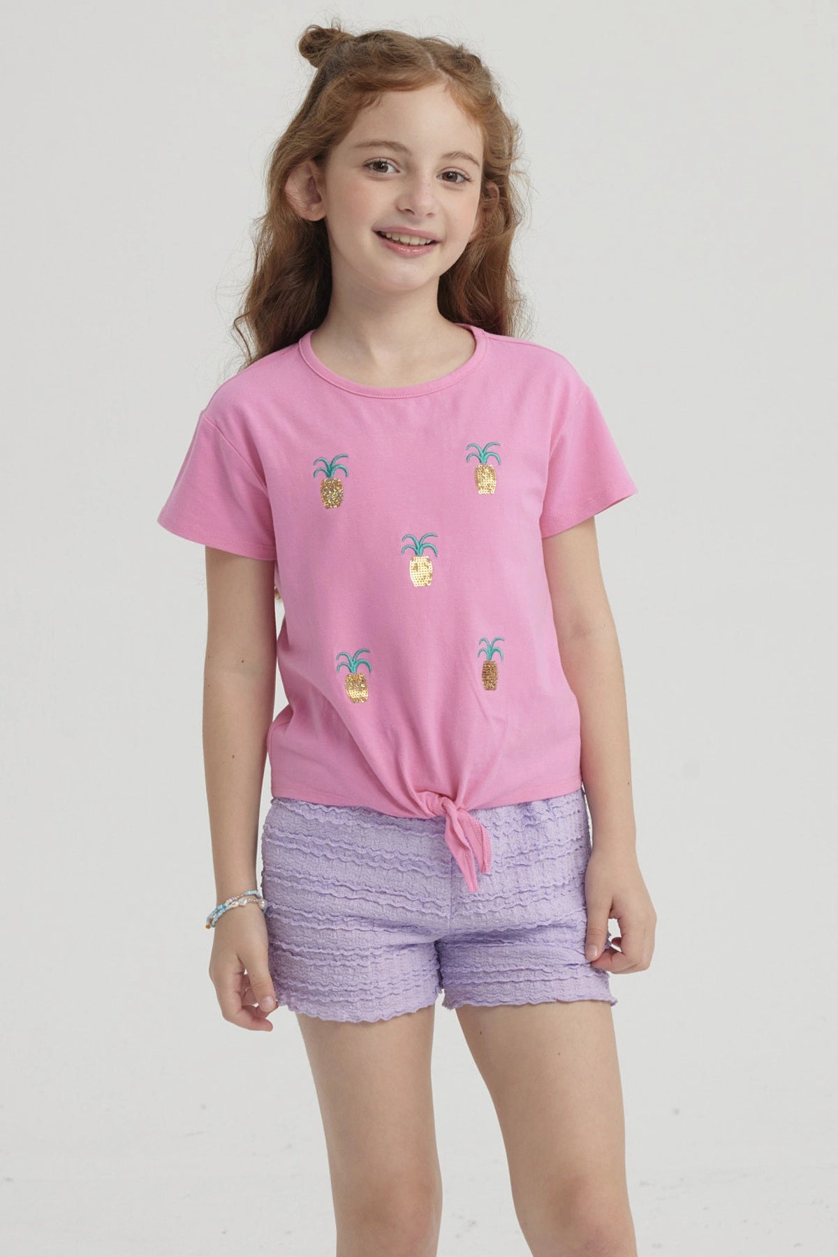 Polera rosada para niña con nudo al frente y estampado de piñas doradas pequeñas distribuidas por toda la prenda