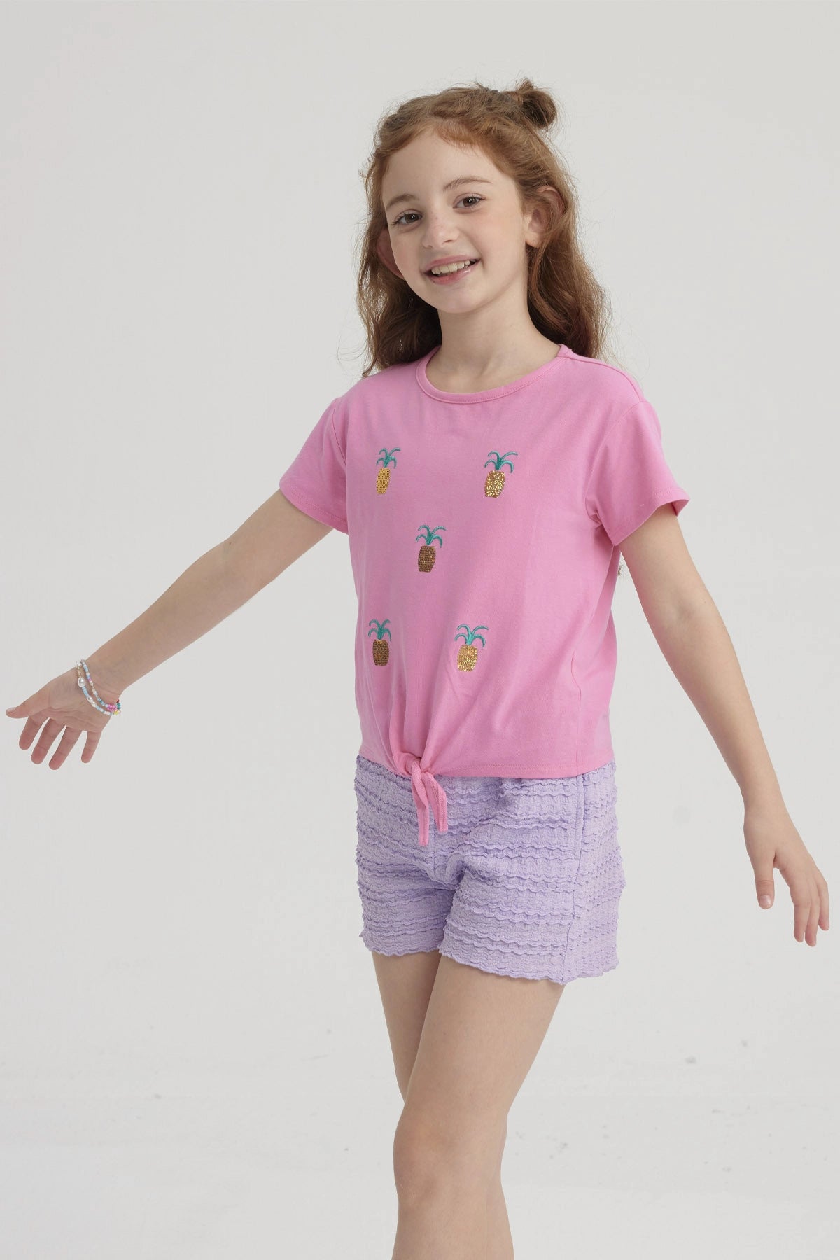 Polera rosada para niña con nudo al frente y estampado de piñas doradas pequeñas distribuidas por toda la prenda