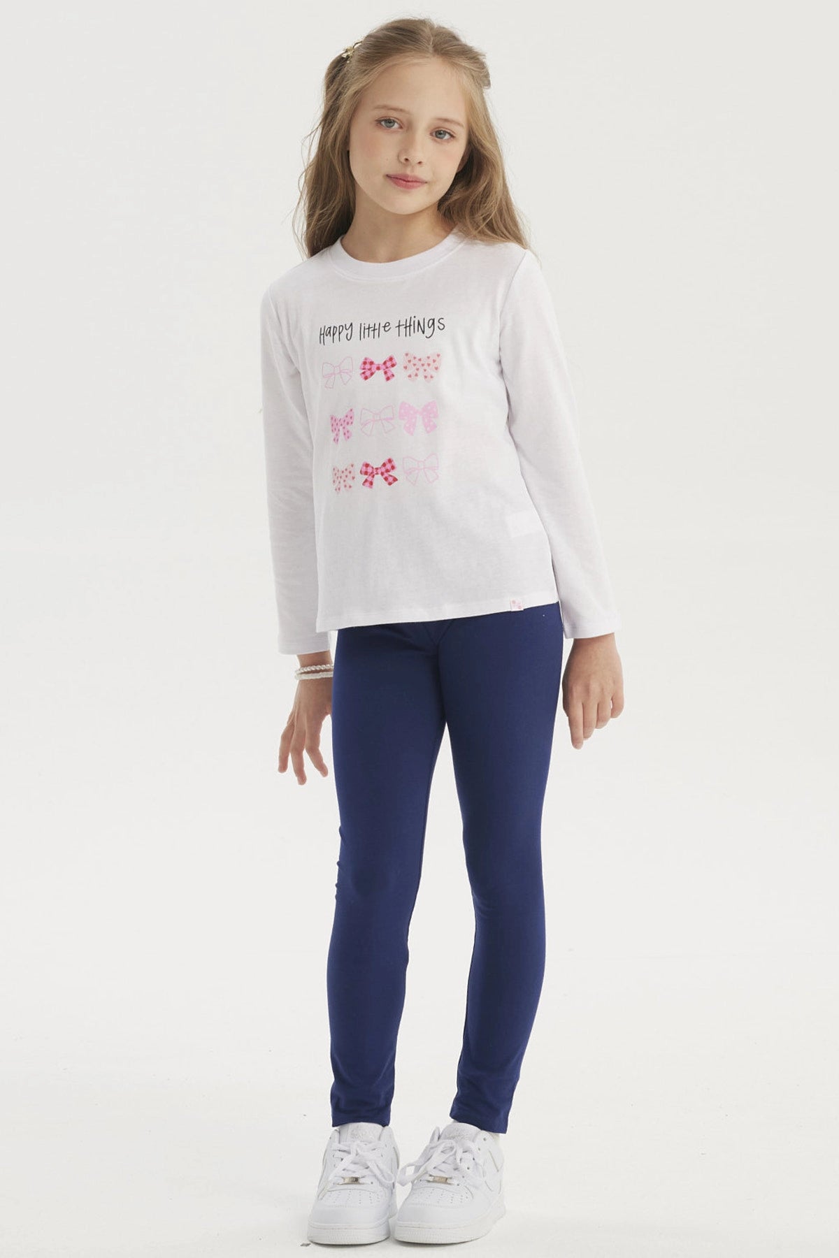 Polera blanca de niña con estampado de lazos en colores rosados y rojos en el centro con texto Happy little things