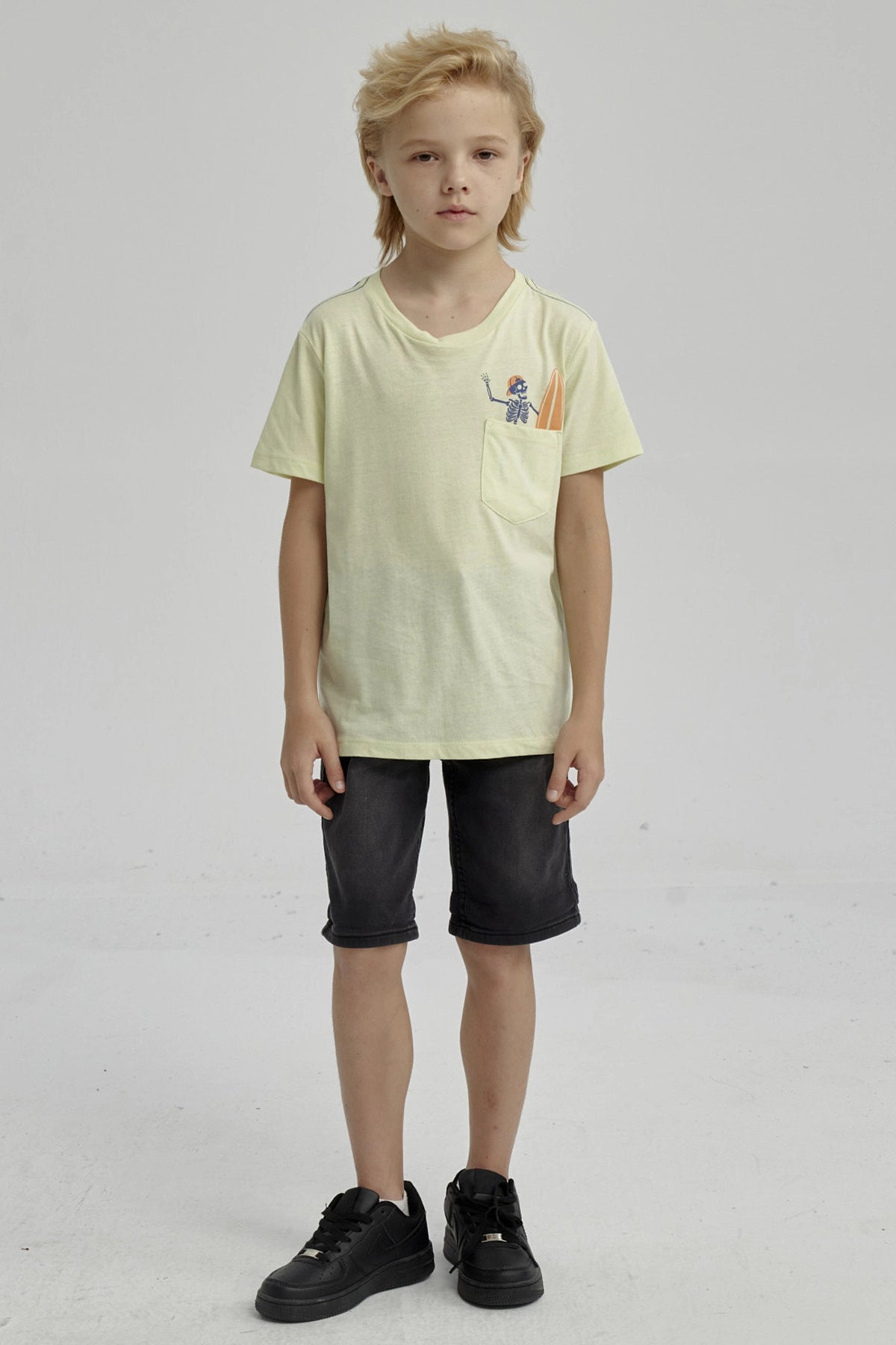 Polera amarilla para niño con bolsillo y estampa de esqueleto surfeando en estilo gráfico pequeño sobre el pecho