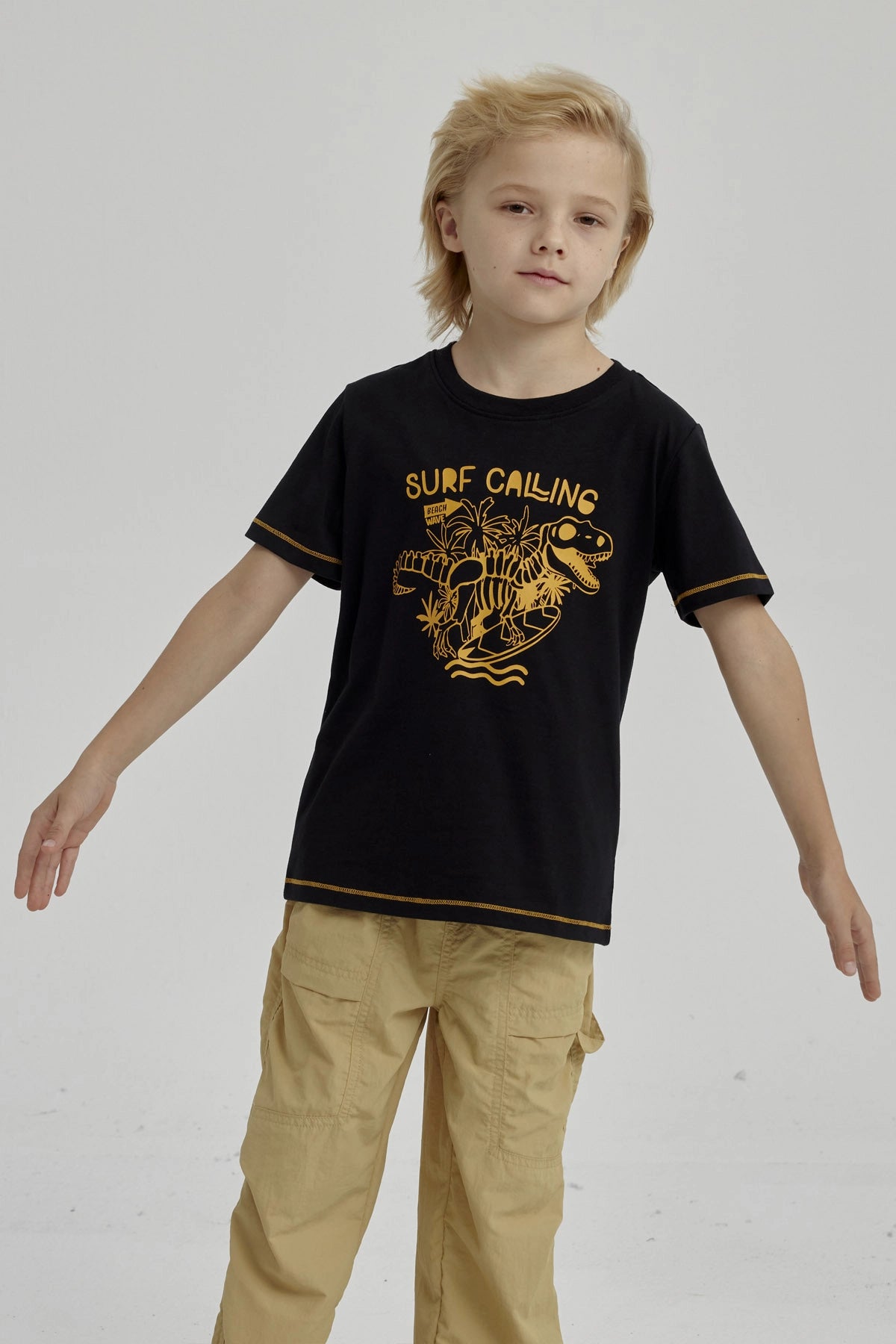 Polera negra para niño con diseño de dinosaurio surfeando y texto Surf Calling en el pecho estilo gráfico amarillo