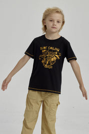 Polera negra para niño con diseño de dinosaurio surfeando y texto Surf Calling en el pecho estilo gráfico amarillo
