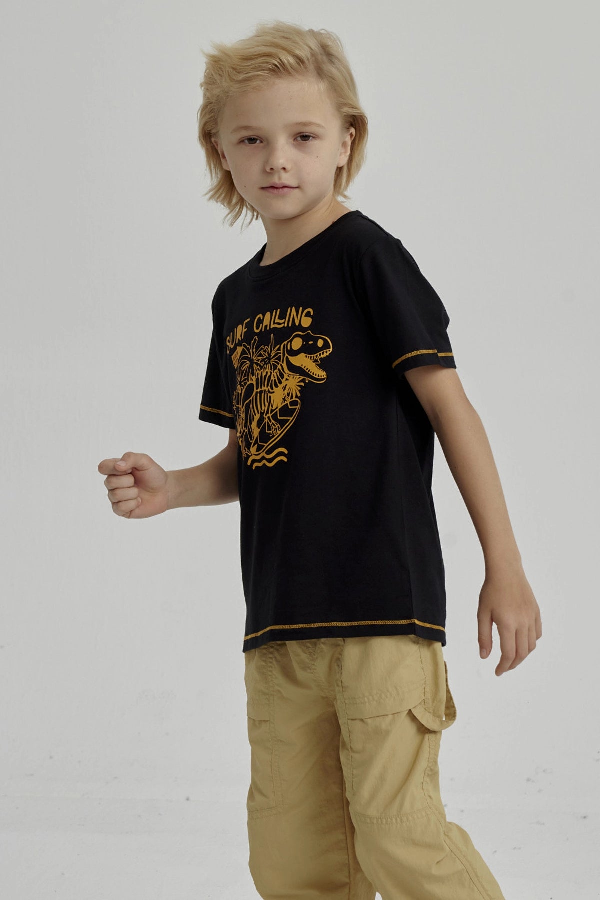 Polera negra para niño con diseño de dinosaurio surfeando y texto Surf Calling en el pecho estilo gráfico amarillo
