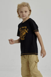 Polera negra para niño con diseño de dinosaurio surfeando y texto Surf Calling en el pecho estilo gráfico amarillo