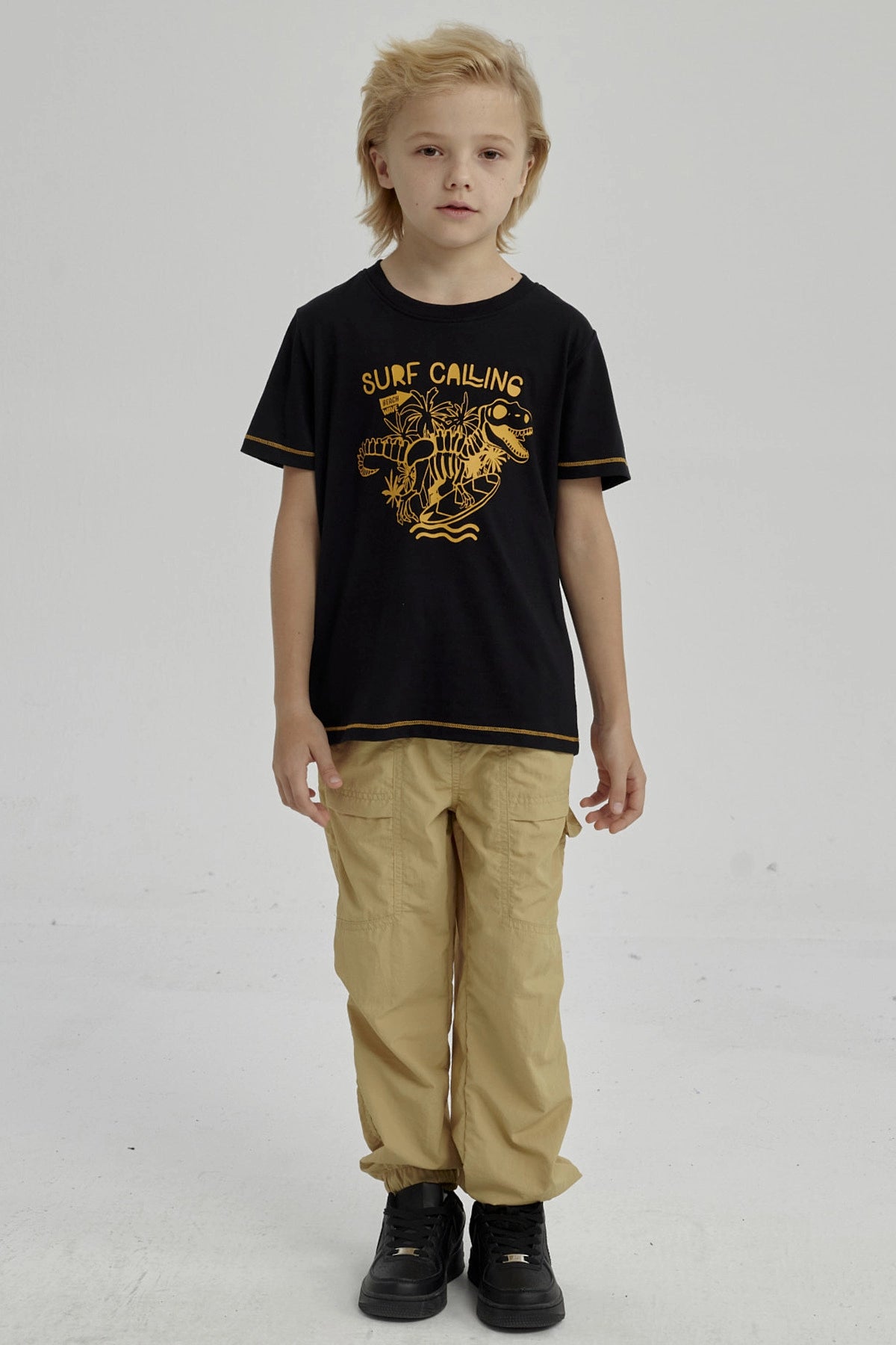 Polera negra para niño con diseño de dinosaurio surfeando y texto Surf Calling en el pecho estilo gráfico amarillo