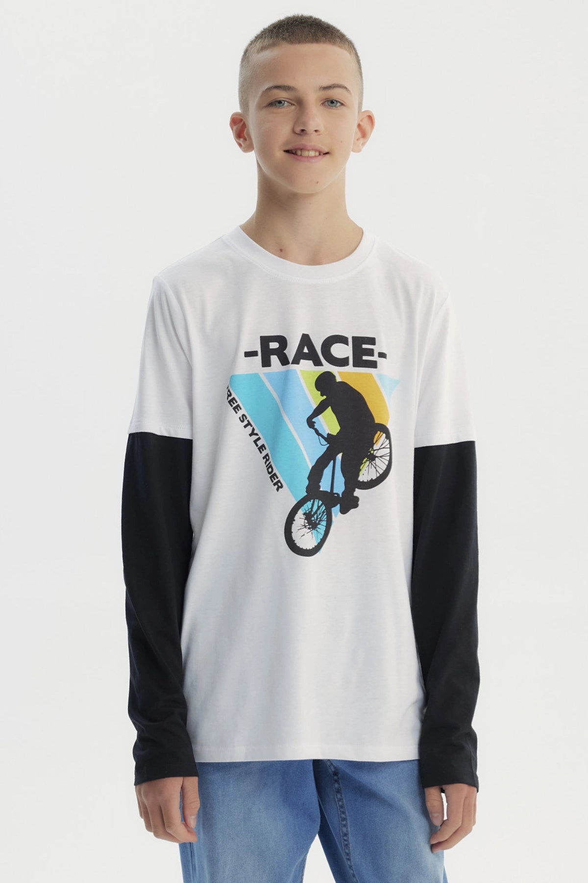Polera niño blanca con mangas largas negras y estampa de ciclista freestyle en el centro sobre fondo de franjas azules y amarillas