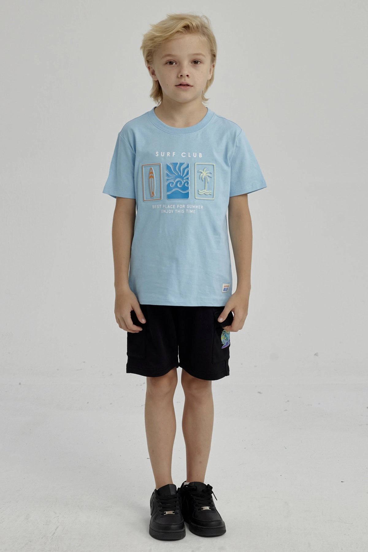 Polera celeste de niño con estampado frontal de surf club con tabla de surf ola y palmera diseño gráfico pequeño