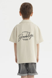 Camisa niño color crudo con cuello abierto y texto Brooklyn en el pecho estilo casual sin estampa adicional