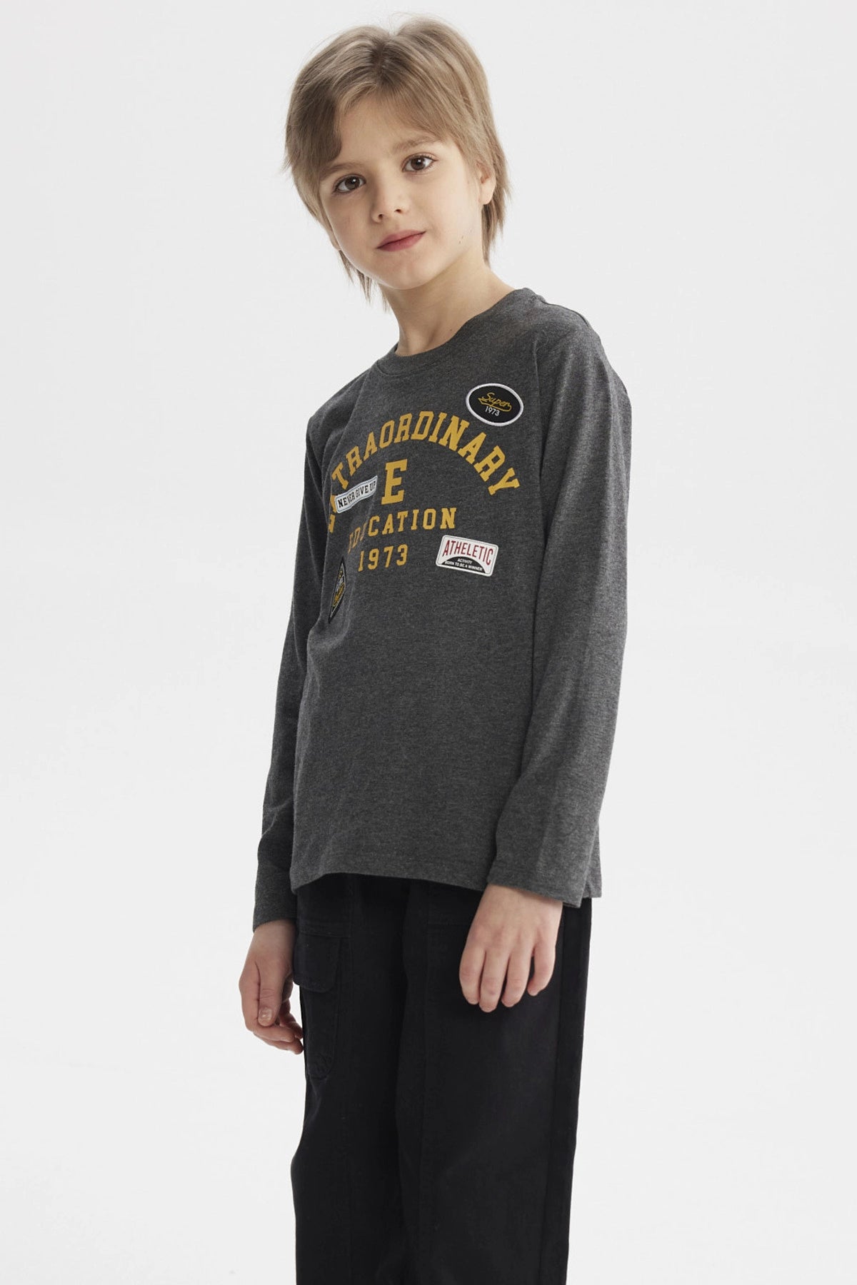 Polera niño gris con parches grandes en el pecho estilo universitario letras amarillas que dicen Extraordinary Education 1973