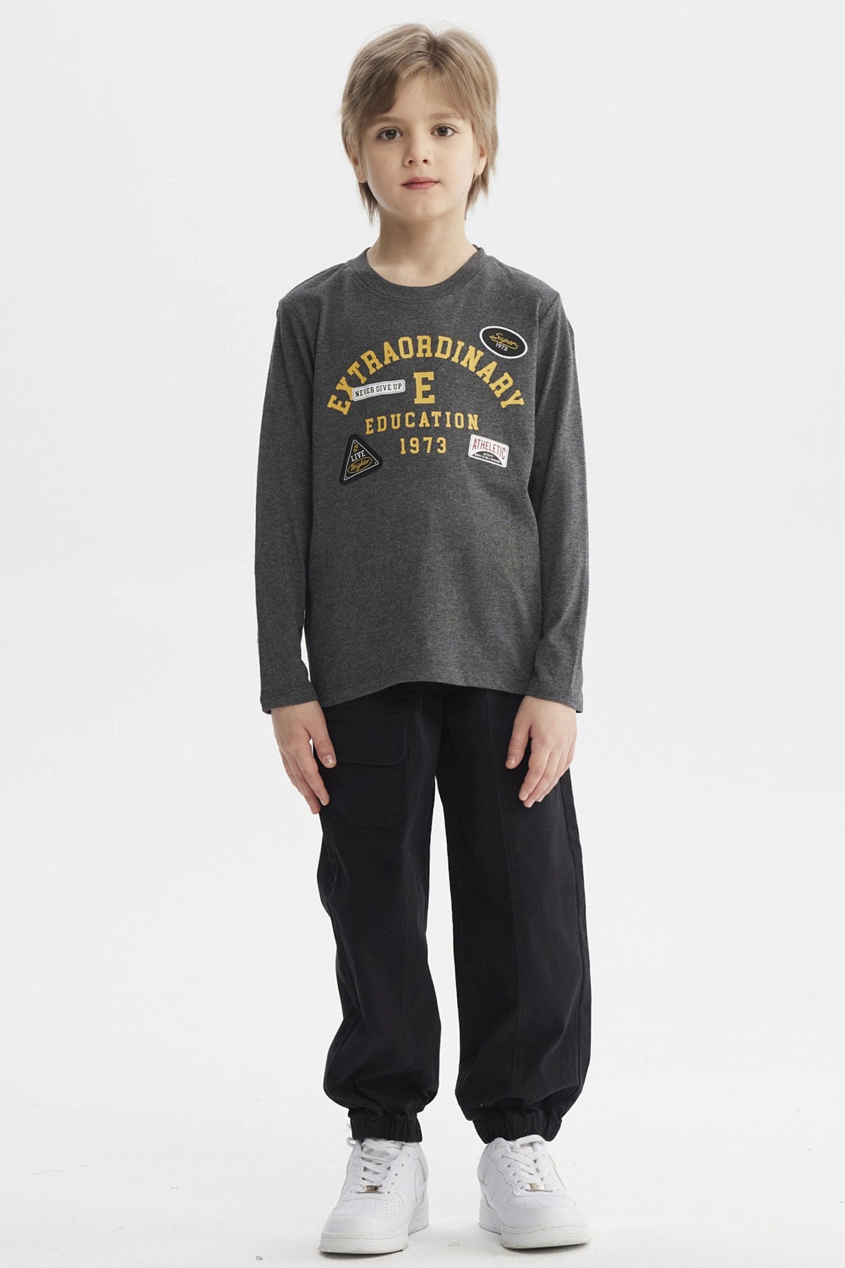 Polera niño gris con parches grandes en el pecho estilo universitario letras amarillas que dicen Extraordinary Education 1973